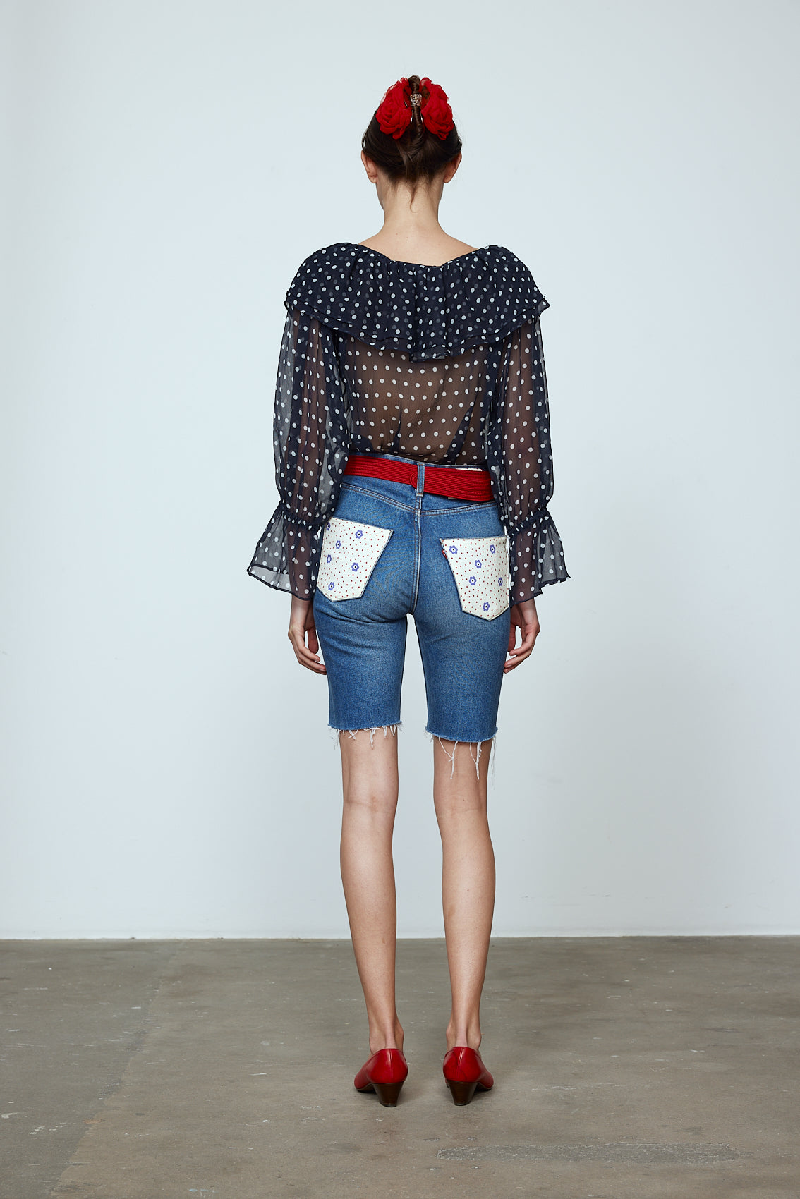 Ursula Blouse in Black Polka Dot Transparent Silk Chiffon. The top features a ruffled boat neckline and sleeves with ruffled long cuffs. Style it with jeans or shorts and a red belt. Material: 100% mulberry silk.