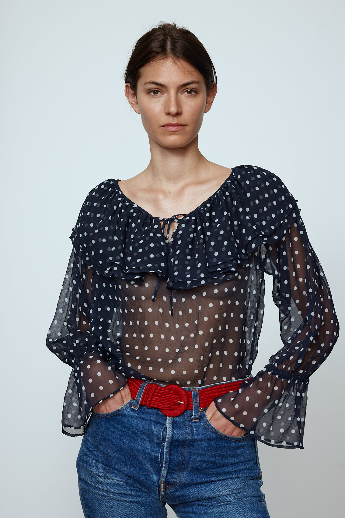 Ursula Blouse in Black Polka Dot Transparent Silk Chiffon. The top features a ruffled boat neckline and sleeves with ruffled long cuffs. Style it with jeans or shorts and a red belt. Material: 100% mulberry silk.