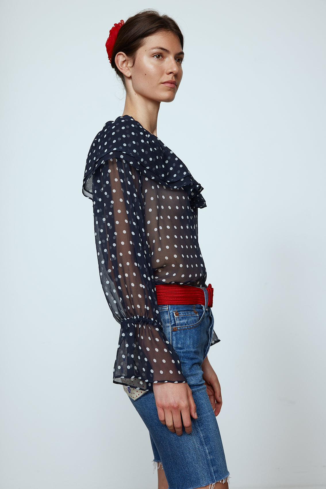Ursula Blouse in Black Polka Dot Transparent Silk Chiffon. The top features a ruffled boat neckline and sleeves with ruffled long cuffs. Style it with jeans or shorts and a red belt. Material: 100% mulberry silk.