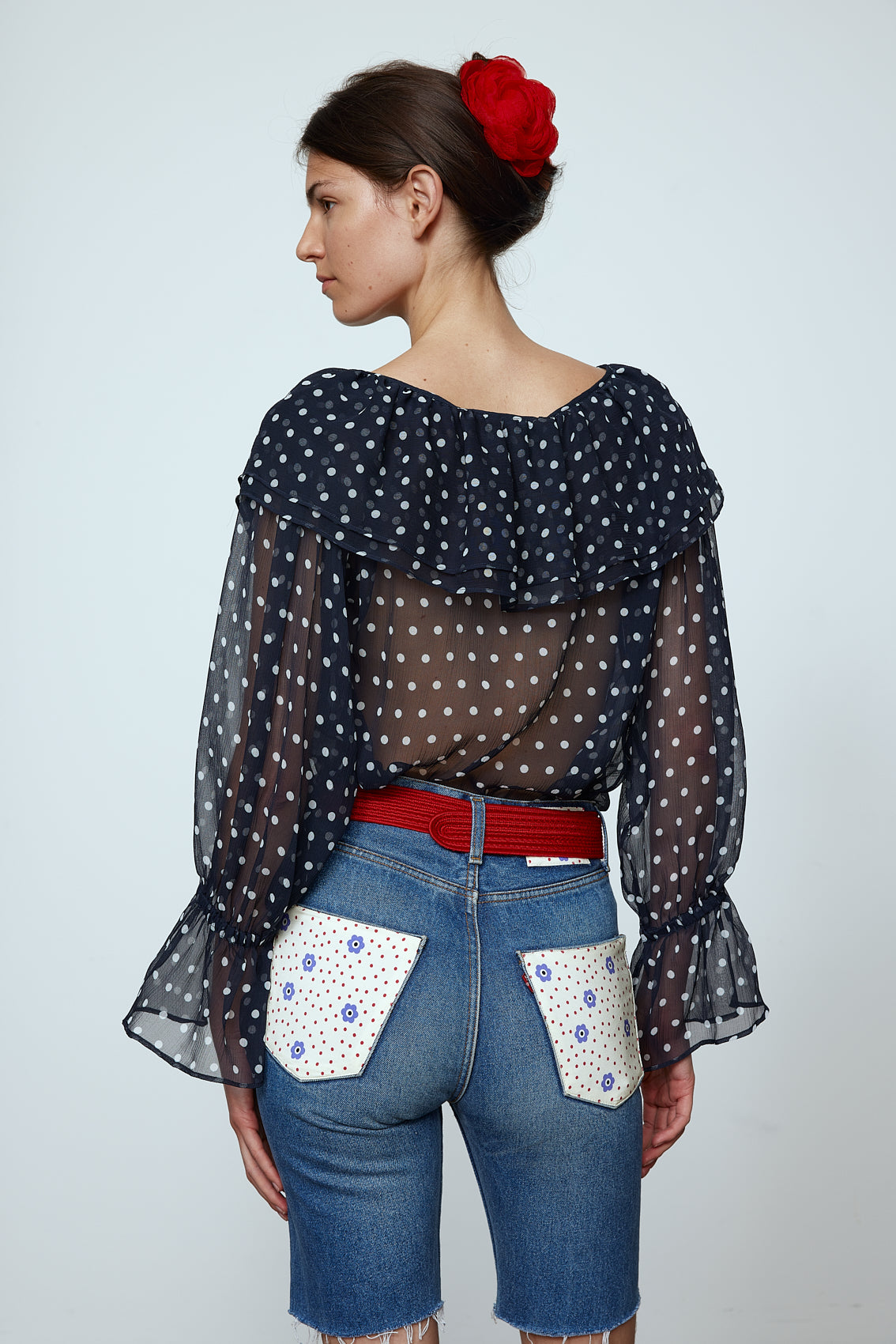 Ursula Blouse in Black Polka Dot Transparent Silk Chiffon. The top features a ruffled boat neckline and sleeves with ruffled long cuffs. Style it with jeans or shorts and a red belt. Material: 100% mulberry silk.